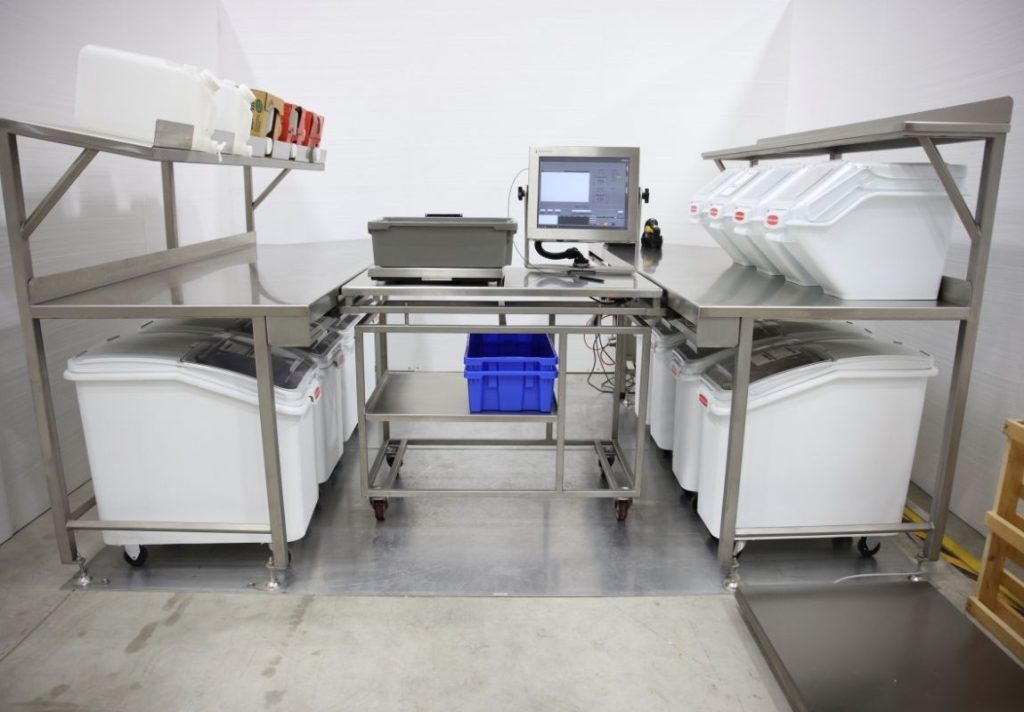 BatchControl Pro 7 | Sterling Systems & Controls Industrial stainless-steel worktable setup with white bins on both sides, a monitor and blue crate in the center, in a clean processing room.