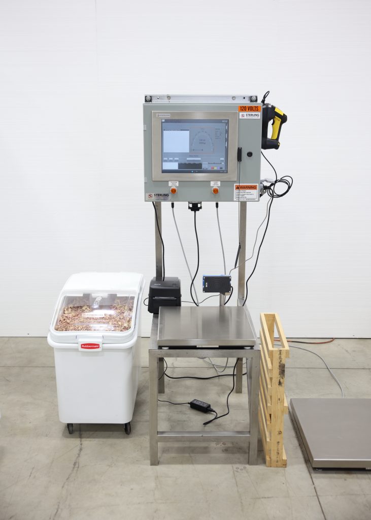 batchcontrol-pro-batching-system-1 | Sterling Systems & Controls Industrial setup with a mounted control panel and touchscreen on a metal frame, wires hanging below, and a yellow-handled tool attached to the side; a weighing table sits below the panel.